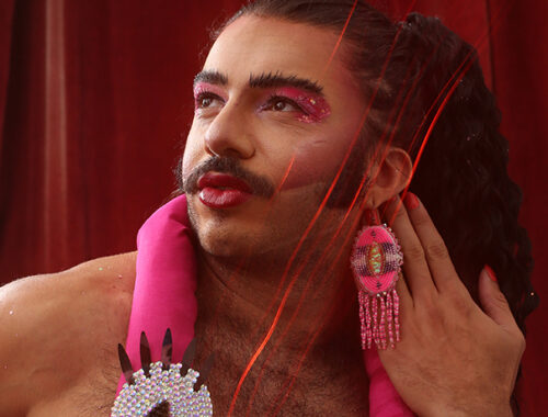 Alex, Glamputee, looks up and to the left side of the picture, pursing lips. Glamputee is in drag makeup, wearing bright pink eyeshadow, blush, and lipstick. They wears long, bright pink dangly beaded earrings, and have a bright pink decorative stuffed flamingo draped around their neck.