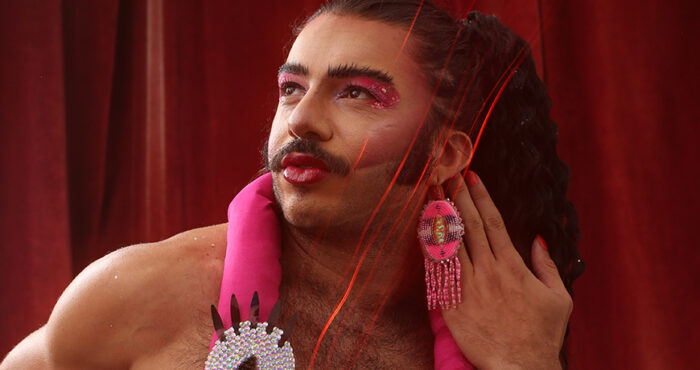 Alex, Glamputee, looks up and to the left side of the picture, pursing lips. Glamputee is in drag makeup, wearing bright pink eyeshadow, blush, and lipstick. They wears long, bright pink dangly beaded earrings, and have a bright pink decorative stuffed flamingo draped around their neck.