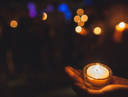 Person holding a small lit candle in their hand, surrounded by darkness with a few twinkling lights in the background