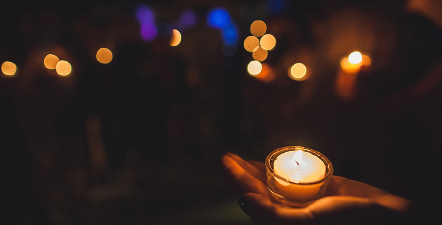 Person holding a small lit candle in their hand, surrounded by darkness with a few twinkling lights in the background