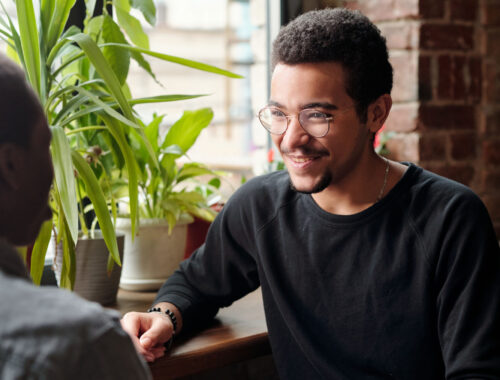 Two young men sitting down at a coffee shop facing each other. Person whose face can be seen is smiling.