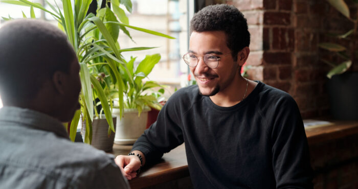 Two young men sitting down at a coffee shop facing each other. Person whose face can be seen is smiling.