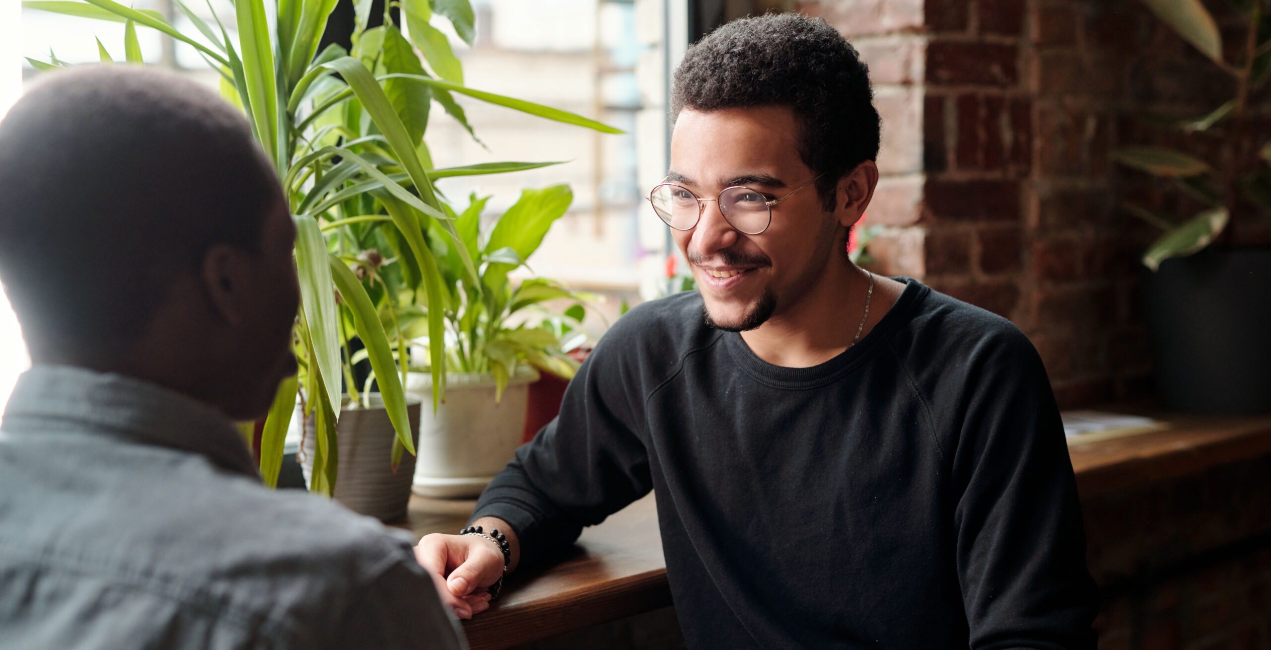 Two young men sitting down at a coffee shop facing each other. Person whose face can be seen is smiling.