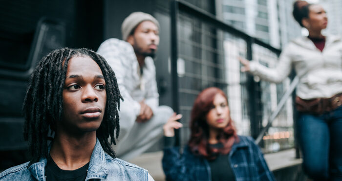 Group of four diverse young people standing by a metal fence, looking into the distance with serious expressions on their faces.