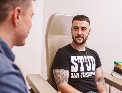 Patient wearing a black T shirt sits in a chair at SFAF's Magnet clinic, accompanied by a clinician standing off to the side