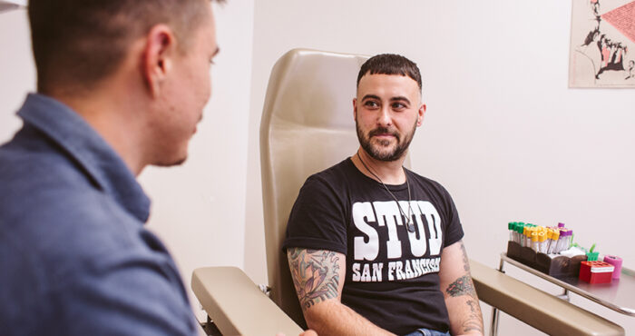 Patient wearing a black T shirt sits in a chair at SFAF's Magnet clinic, accompanied by a clinician standing off to the side