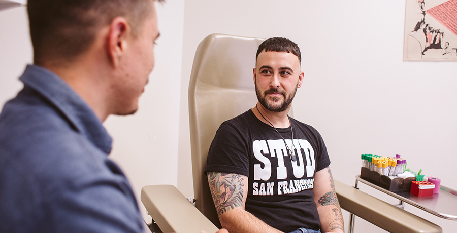 Patient wearing a black T shirt sits in a chair at SFAF's Magnet clinic, accompanied by a clinician standing off to the side