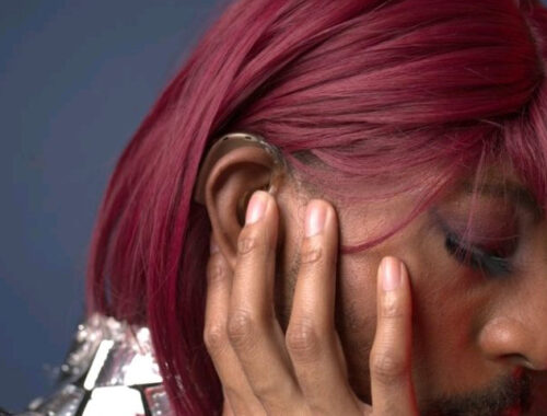 Kevin Abrams, aka Iman., side view, looking down and to the left, away from the camera. Wearing a shiny silver top, and dark red wig, colorful eye makeup, has a hand cupping a cheek and a hearing aid over the ear is visible.