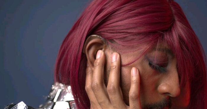 Kevin Abrams, aka Iman., side view, looking down and to the left, away from the camera. Wearing a shiny silver top, and dark red wig, colorful eye makeup, has a hand cupping a cheek and a hearing aid over the ear is visible.