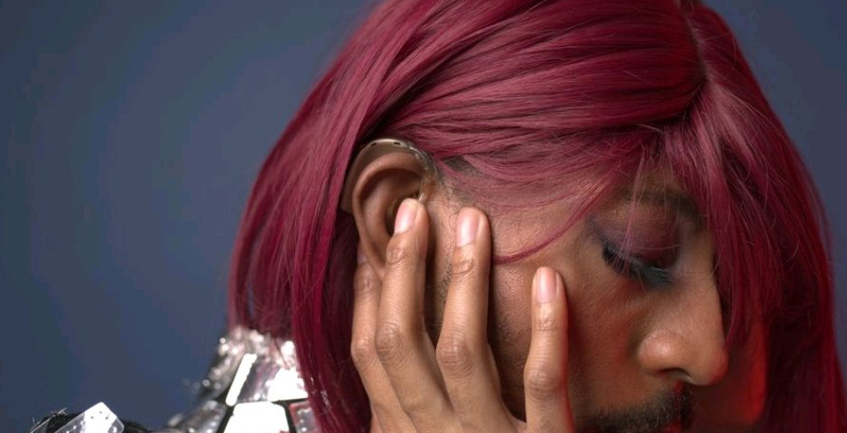 Kevin Abrams, aka Iman., side view, looking down and to the left, away from the camera. Wearing a shiny silver top, and dark red wig, colorful eye makeup, has a hand cupping a cheek and a hearing aid over the ear is visible.